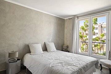 A neutral-toned bedroom with textured grey walls, a made bed, nightstands with lamps, and glass balcony doors showing trees.