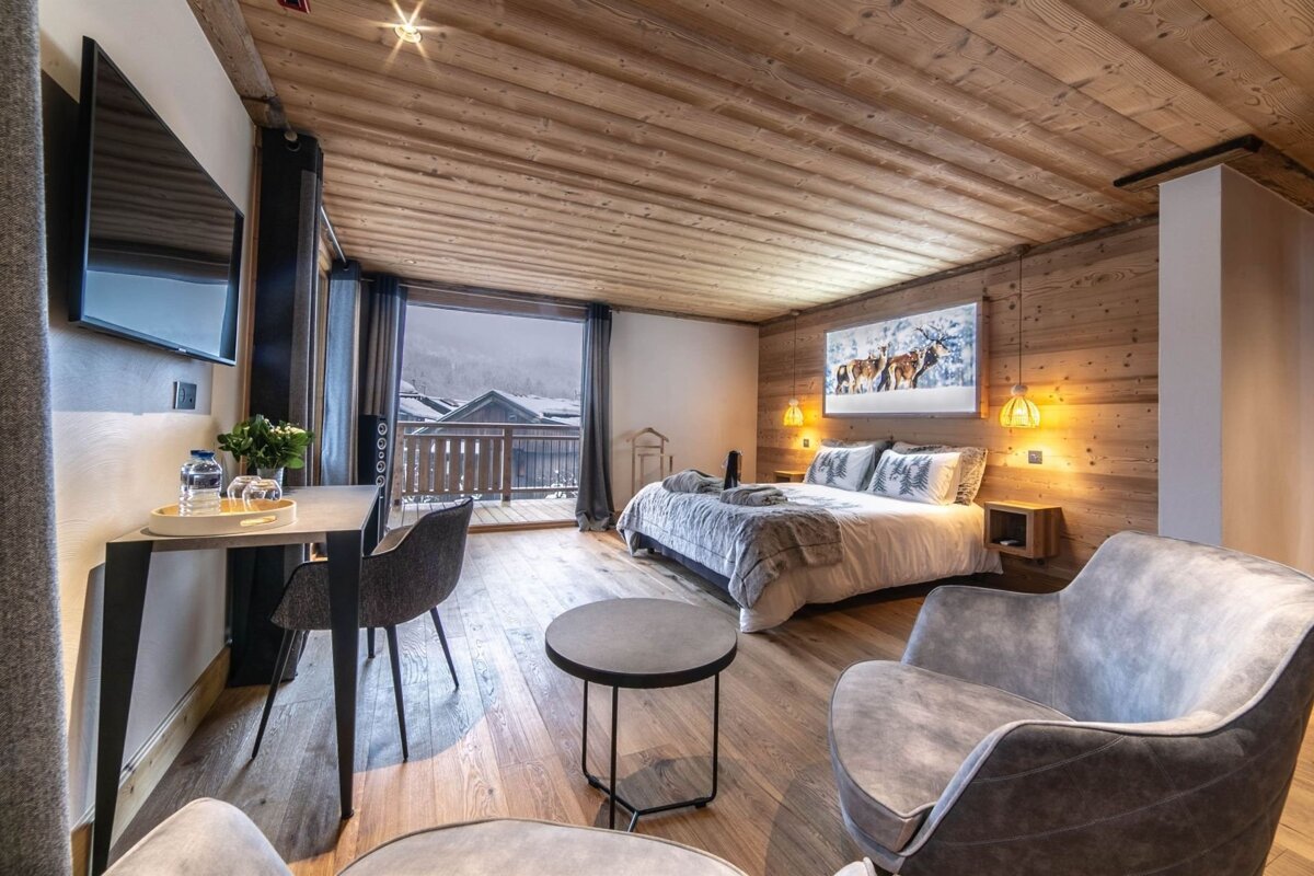 Cozy chalet bedroom featuring warm wood decor, modern furniture, and a balcony overlooking a snowy village scene.