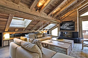 A warm, rustic attic living room featuring exposed wooden beams, skylights, comfy couches, a fireplace with a TV, and modern coffee tables.