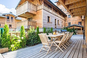 A wooden dining table and chairs on a spacious deck outside rustic alpine chalets, bordered by green plants.