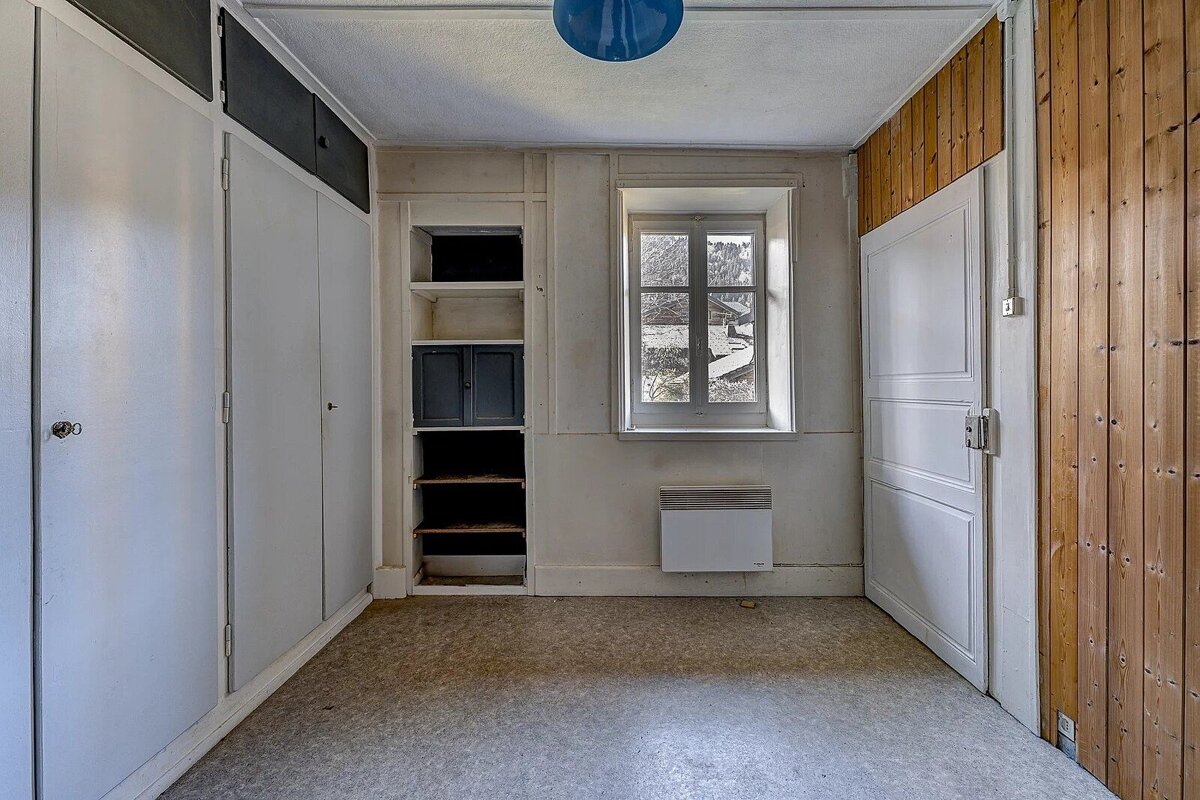 An empty room with built-in white wardrobes, shelving, a window with an outdoor view, a white door, wood paneling, and a blue ceiling light.