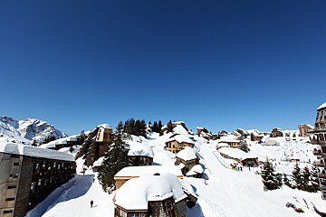 A sprawling snow-covered mountain ski resort under a bright blue sky, featuring numerous unique chalets, pine trees, and active ski slopes.