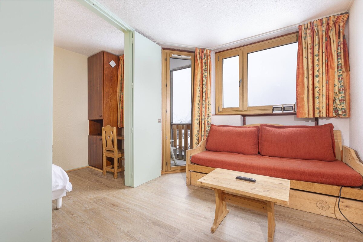 A cozy studio apartment with a red sofa, wooden table, and a balcony view of a snowy landscape, featuring an adjacent work/bedroom nook.
