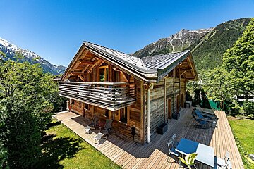 Charming wooden chalet with a spacious deck, surrounded by lush greenery and majestic snow-capped mountains under a clear blue sky.