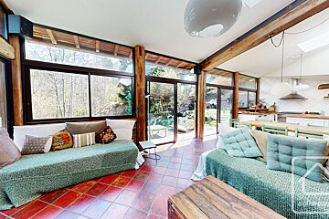 A sunlit rustic living room with tiled floors, large windows, wooden pillars, and two green-covered sofas.