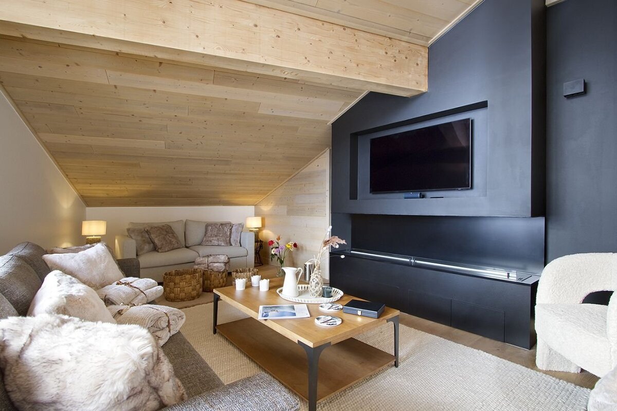 A warm, modern attic living room featuring a sloped wooden ceiling, sleek dark fireplace with TV, and plush seating around a coffee table.
