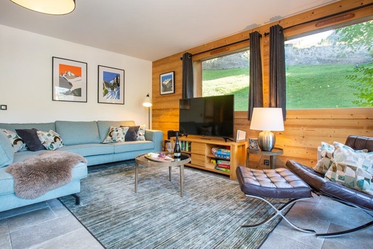 A cozy chalet living room with a blue sofa, wood-paneled walls, a leather lounge chair, and a large window overlooking a grassy hill.