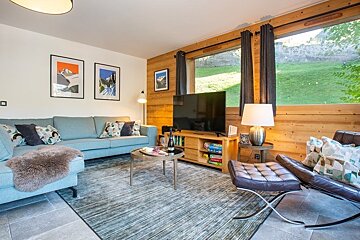 A cozy chalet living room with a blue sofa, wood-paneled walls, a leather lounge chair, and a large window overlooking a grassy hill.