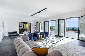 A modern, bright living room features stylish grey and blue furniture, a glass coffee table, and large glass doors opening to a scenic terrace.