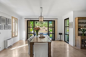A bright dining room featuring a long wooden table, white upholstered chairs, tile floors, and large glass doors looking onto a garden.