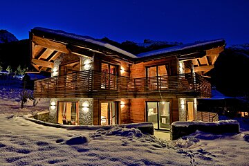 An inviting, warmly lit wooden chalet stands in fresh snow at night under a dark blue sky, nestled in a mountain setting.