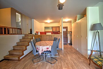 Cozy apartment interior with a dining table, grey chairs, wooden stairs, and a kitchen in the background.