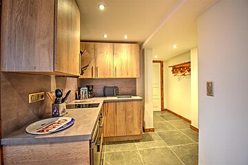 Cozy kitchen with light wood cabinets, gray counters, and tiled floor leading to a hallway with a wooden coat rack.