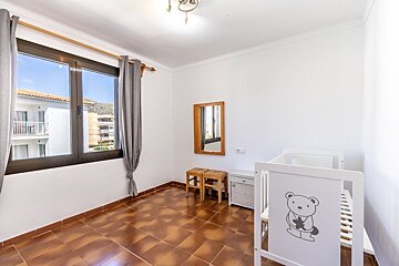 Bright room featuring brown tiled floors, a large window, a wall mirror, stools, and a white baby crib with a cute bear cartoon.