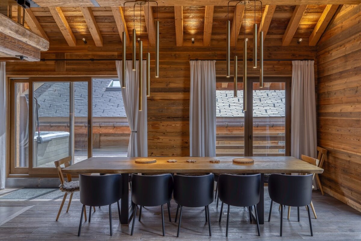 Modern rustic dining room with wooden interior, long table, black chairs, and pendant lights. Sliding doors overlook a hot tub.