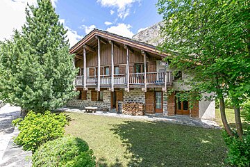A rustic wood and stone chalet with balconies, surrounded by lush green trees and a lawn, set against mountains under a blue, cloudy sky.