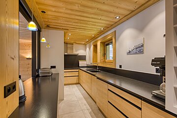 A modern kitchen with a warm wood-paneled ceiling, light wooden cabinets, and dark countertops. Appliances like a toaster and coffee maker are visible.