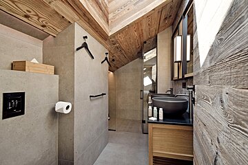 Modern bathroom with a sloped wooden ceiling, concrete walls, double grey vessel sinks on a wooden vanity, and a glass shower.