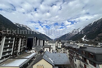 Mountain valley town with buildings, snowy peaks, and a cloudy blue sky.