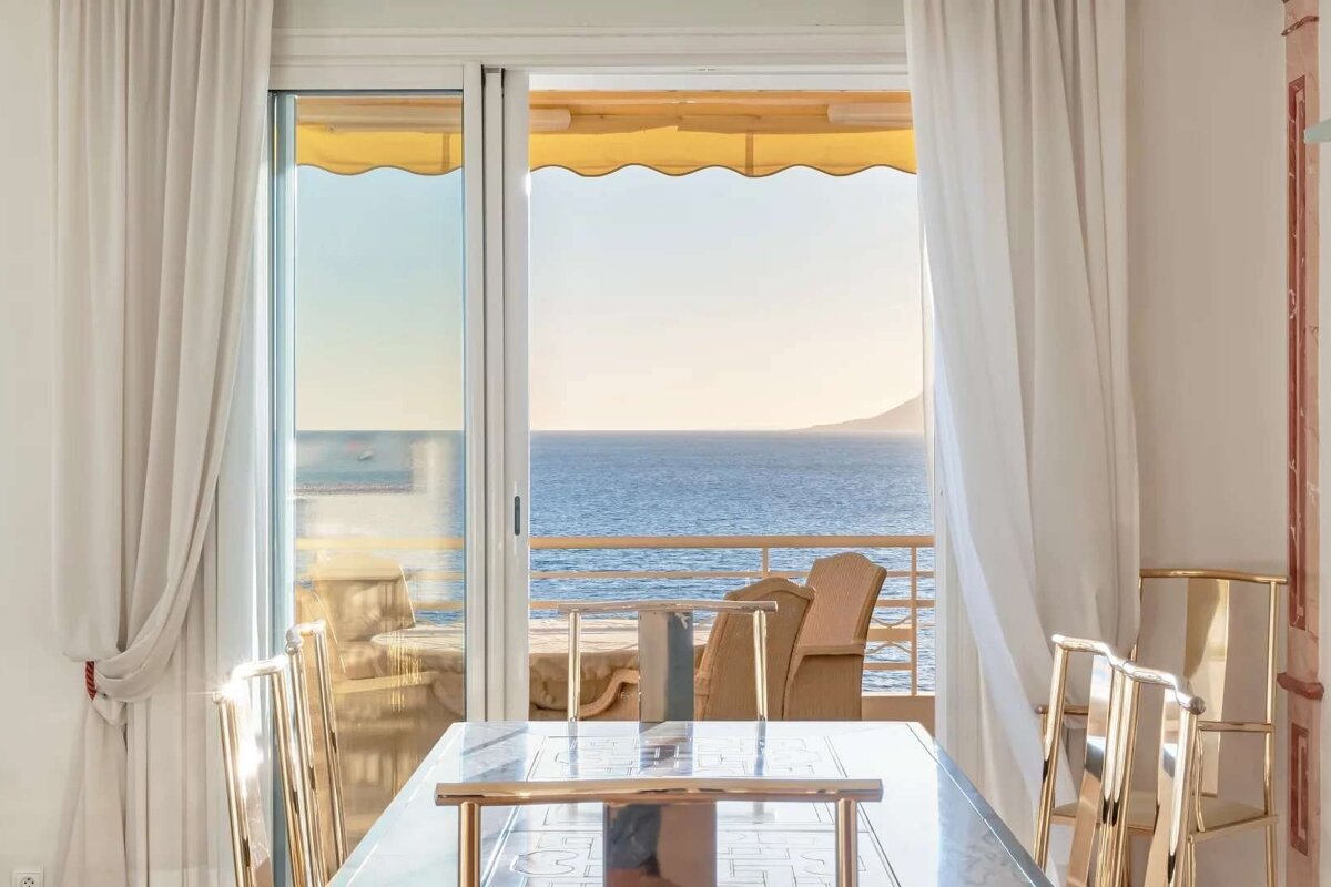 A bright room with white curtains and a dining table, featuring a balcony with a yellow awning and a stunning ocean view stretching to a distant coast.