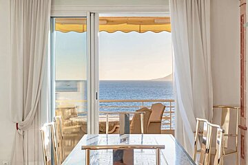 A bright room with white curtains and a dining table, featuring a balcony with a yellow awning and a stunning ocean view stretching to a distant coast.