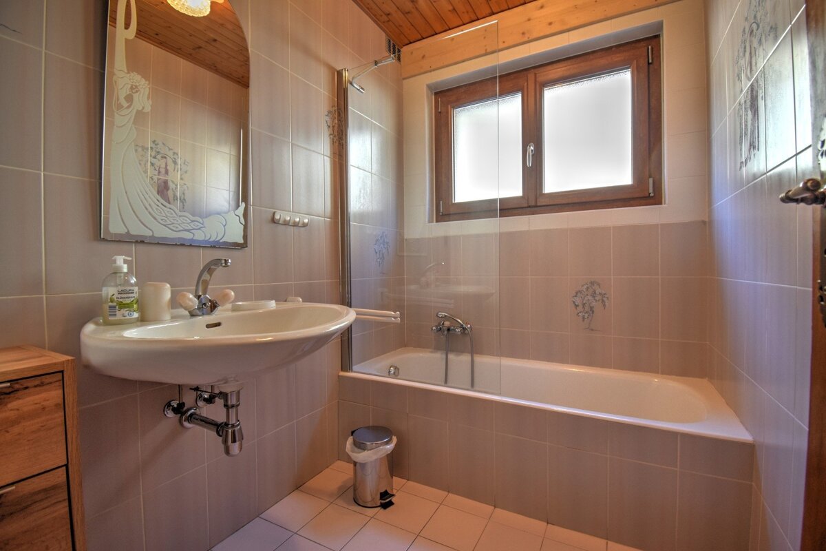 A cozy bathroom with a sink, ornate mirror, bathtub with shower screen, and a window. It features tiled walls, floor, and a wooden ceiling.