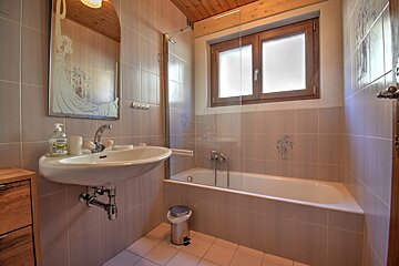 A beige-tiled bathroom with a decorative mirror above a sink, a bathtub with a glass screen, a wooden ceiling, and a window providing natural light.