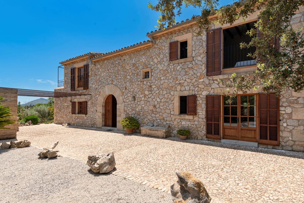 A traditional stone house with wooden shutters and a large arched doorway, set on a mixed gravel and paved courtyard under a bright blue sky.