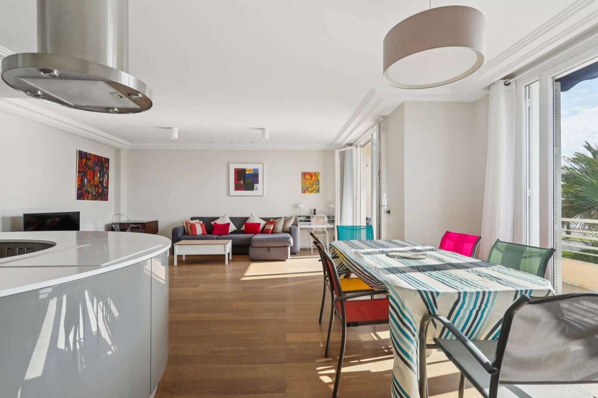 Bright, modern open-plan living space featuring a grey kitchen island, dining table with colorful chairs, a sofa, and abstract art on hardwood floors.