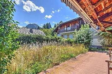 A rustic wooden porch overlooks overgrown grass, a wooden chalet-style house, and another building with a mountain in the background under a blue sky.