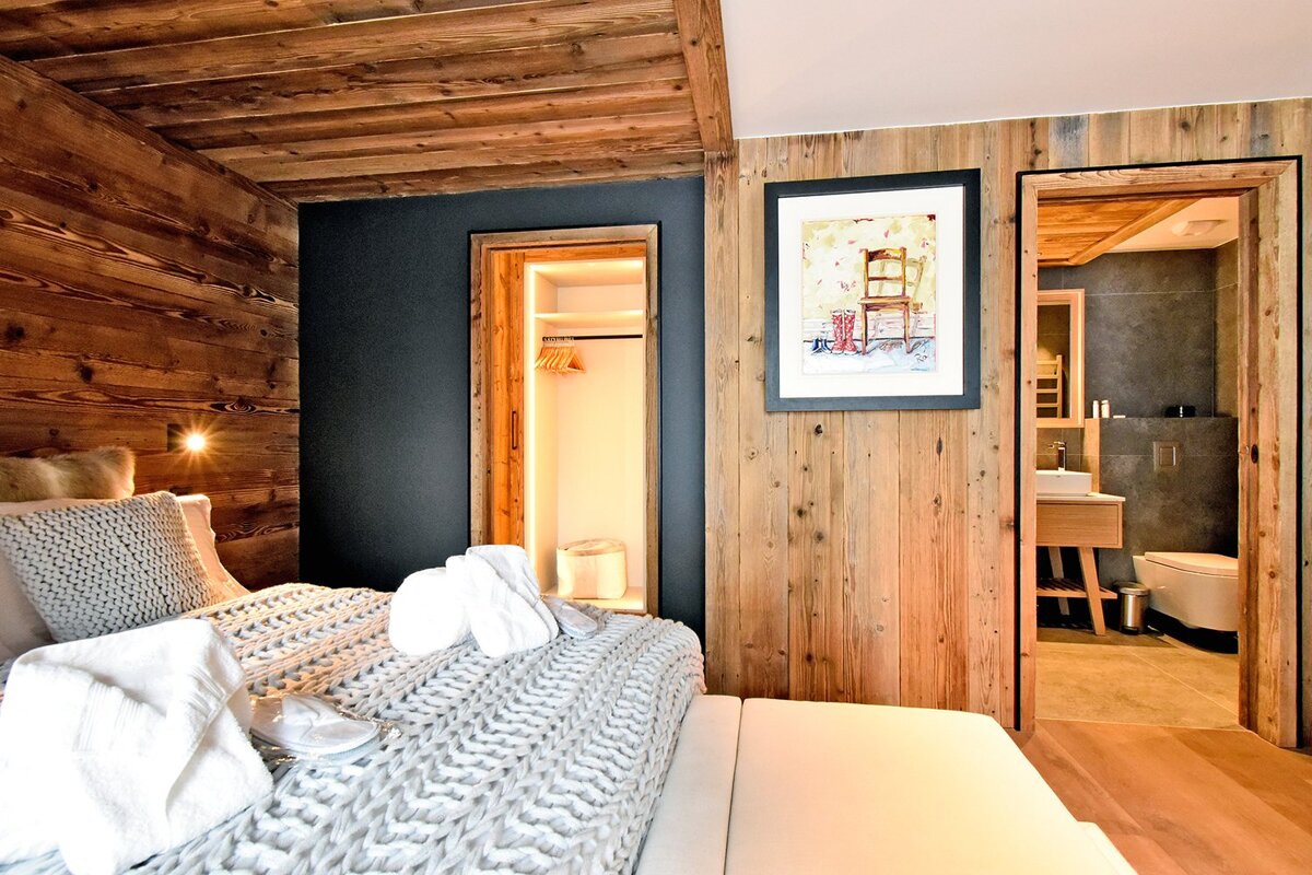 A rustic-chic bedroom with wooden walls & ceiling, cozy bed with knit throw, robes, slippers, an open closet, and a modern bathroom view.