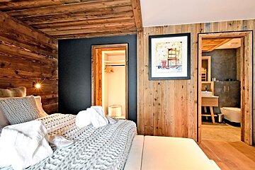 A rustic-chic bedroom with wooden walls & ceiling, cozy bed with knit throw, robes, slippers, an open closet, and a modern bathroom view.