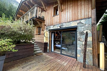 Rustic wooden chalet with a stone accent wall, sliding glass door opening to a bedroom, and a wooden deck surrounded by greenery.