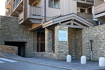 Entrance of the Carré Blanc building featuring stone walls, wooden chalet-style upper levels, and a sign on a pillar.