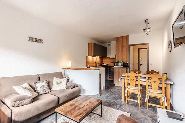 A cozy open-plan living room and kitchen with a brown sofa, wooden coffee table, dining area with heart-cutout chairs, and a modern wooden kitchenette.