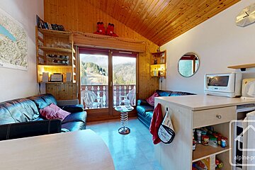 Cozy chalet living space with wooden ceiling, mountain-view balcony, leather sofas, and a compact kitchen.