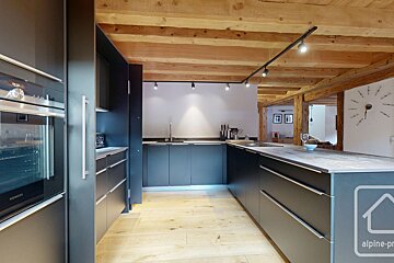 Modern charcoal kitchen with sleek dark cabinets, light wood floor, and rustic exposed wooden beams overhead, illuminated by track lighting.