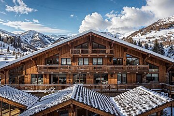 Large wooden alpine chalet with snow-covered roofs, set against majestic snowy mountains under a blue sky.