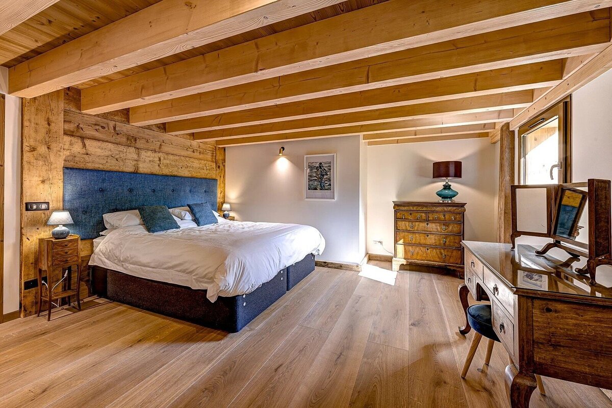 A cozy, rustic bedroom with exposed wooden beams, hardwood floor, a large bed with blue headboard, dresser, and a vanity.