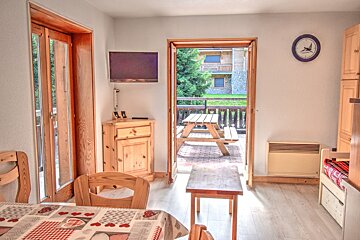A bright room with wooden furniture, a dining table, and a TV. An open door leads to a patio with a picnic table, trees, and buildings visible.