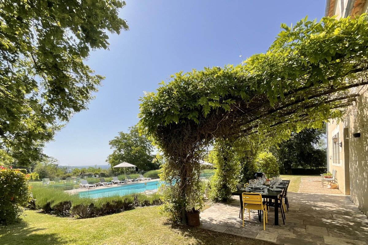A patio with a table and chairs under a pergola overlooking a swimming pool