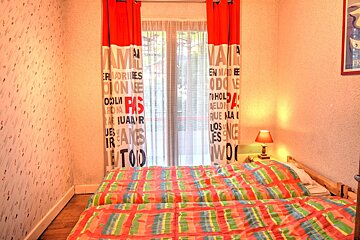 A bright bedroom features two beds with vibrant striped comforters, red and white text-patterned curtains by a window, and light patterned wallpaper.