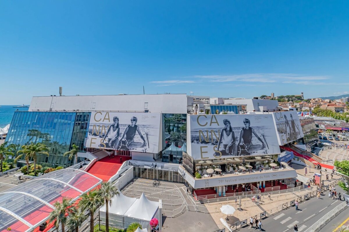 An aerial view of the Palais des Festivals in Cannes decorated with large posters and the iconic red carpet under a bright blue sky.