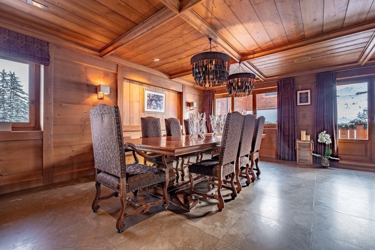 A cozy, wood-paneled dining room features a large wooden table with ornate chairs, two striking chandeliers, and windows revealing a snowy mountain view.