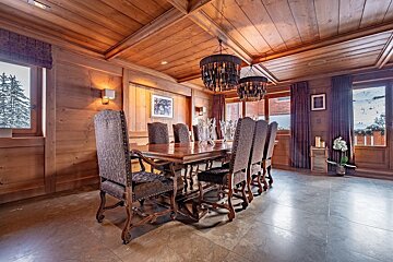 A cozy, wood-paneled dining room features a large wooden table with ornate chairs, two striking chandeliers, and windows revealing a snowy mountain view.