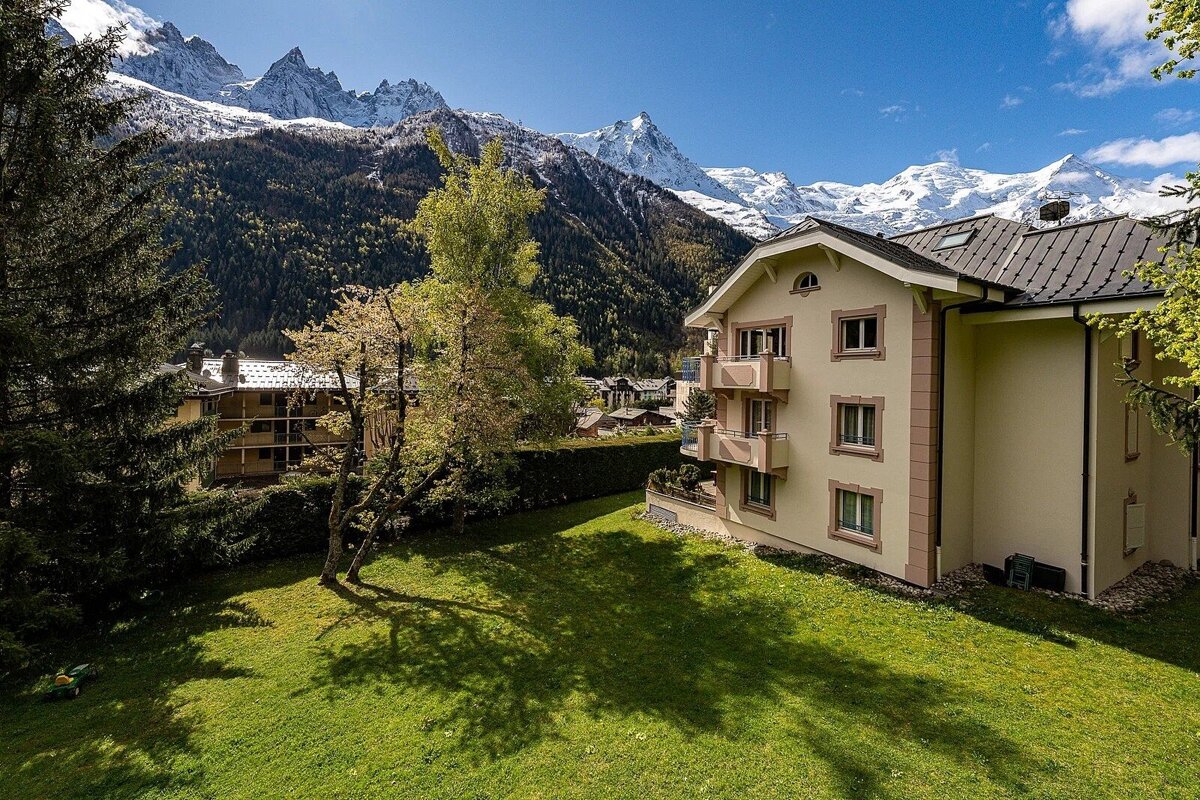 A beige building with balconies overlooks a green lawn and trees, set against a stunning backdrop of snow-capped mountains under a clear sky.