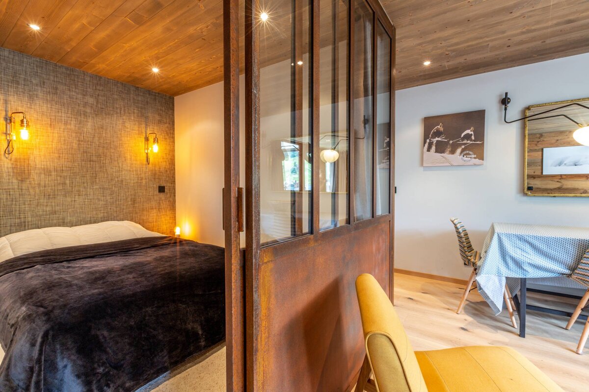 A rustic modern interior featuring a cozy bedroom with wood ceiling & industrial lighting, separated from a dining area by a rusty metal and glass partition.