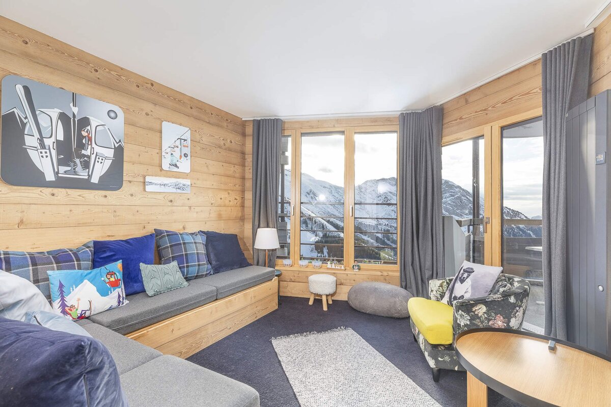Cozy, wood-paneled ski chalet living room with a large window overlooking snowy mountains, dark carpet, and ski-themed art and pillows.