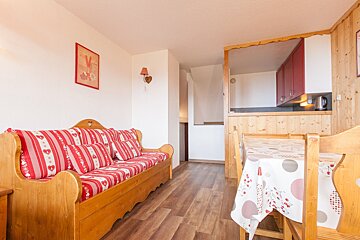 Cozy, rustic studio with a wooden sofa, red striped cushions, dining table with patterned cloth, and a small kitchen with red cabinets.