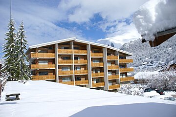 A modern building with wooden balconies in a snow-covered mountain landscape, with pine trees, distant peaks, and a blue sky.